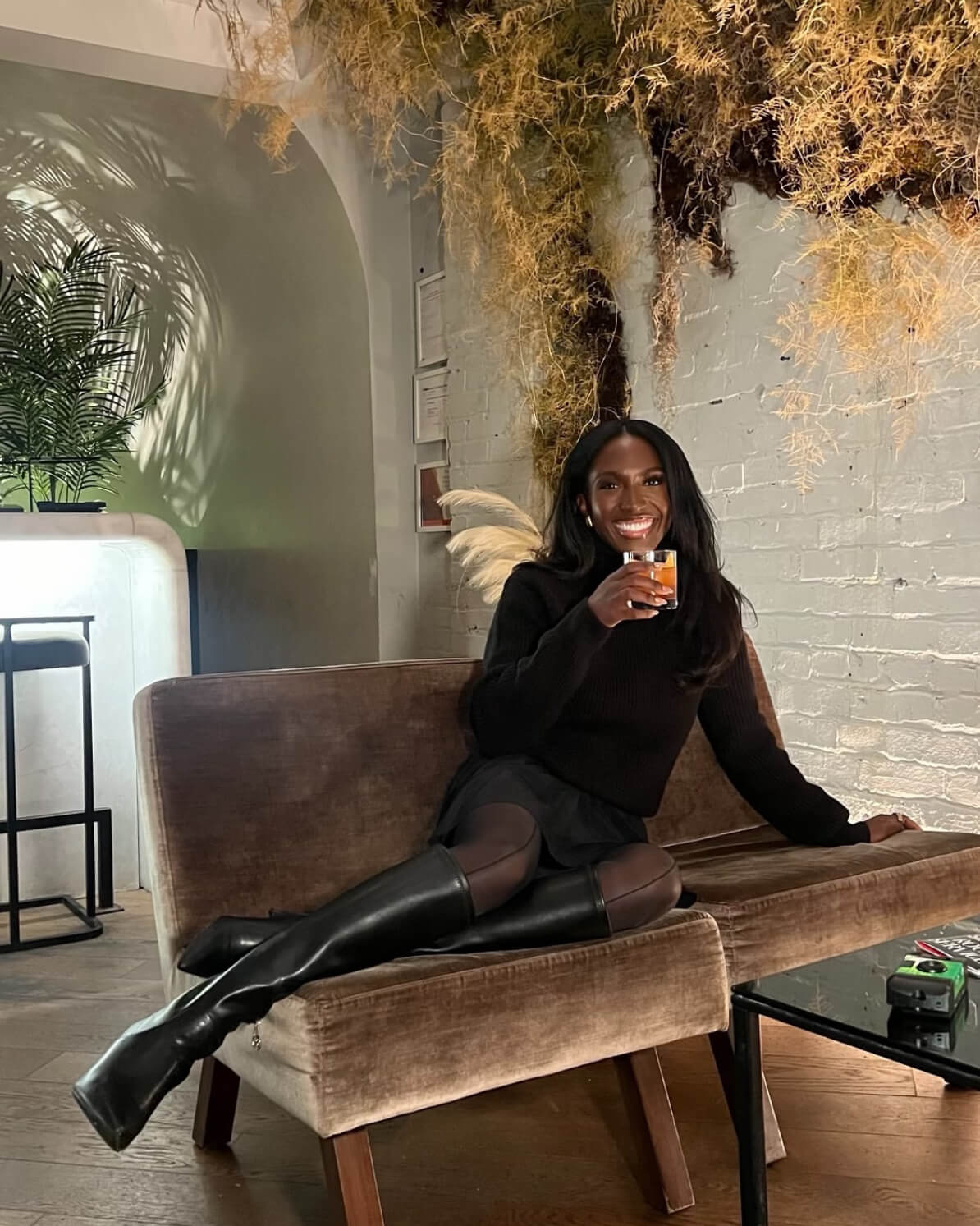Woman sitting on a brown sofa holding a drink in a modern indoor setting with plants.